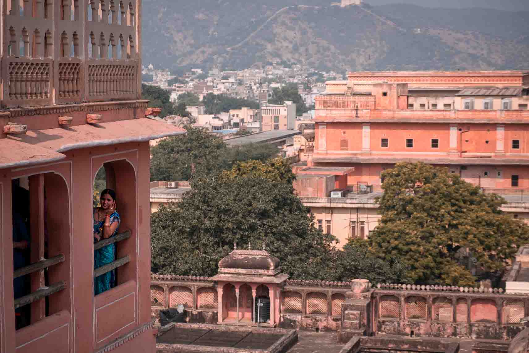 A woman in a blue sari at a pink palace window, the rooftops of Jaipur rising behind her