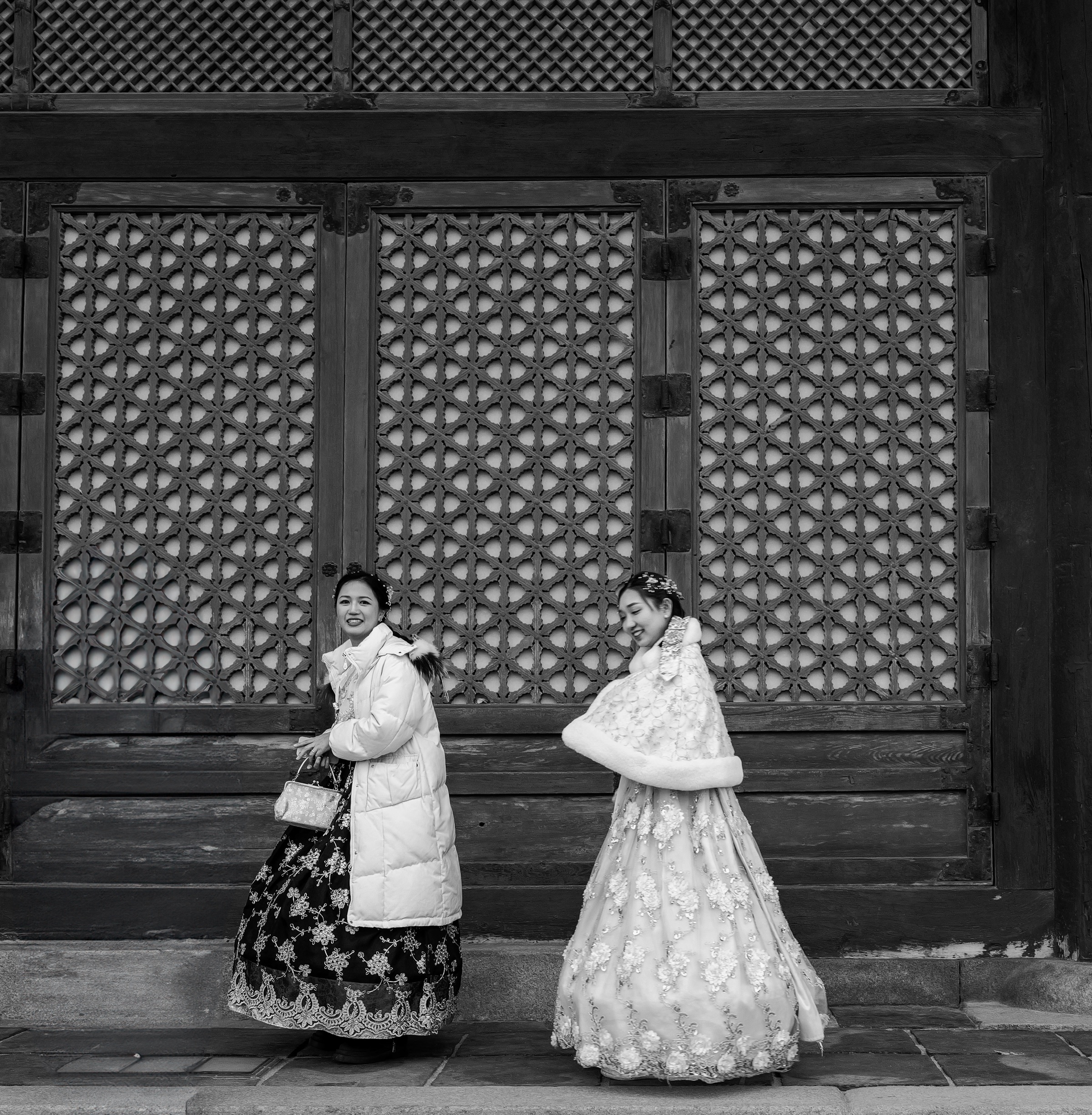 Two women in hanbok and winter coats passing the latticed doors of a palace hall