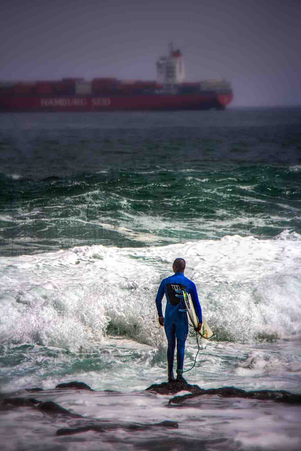 Surfer in wetsuit walking into the surf with a container ship looming on the horizon