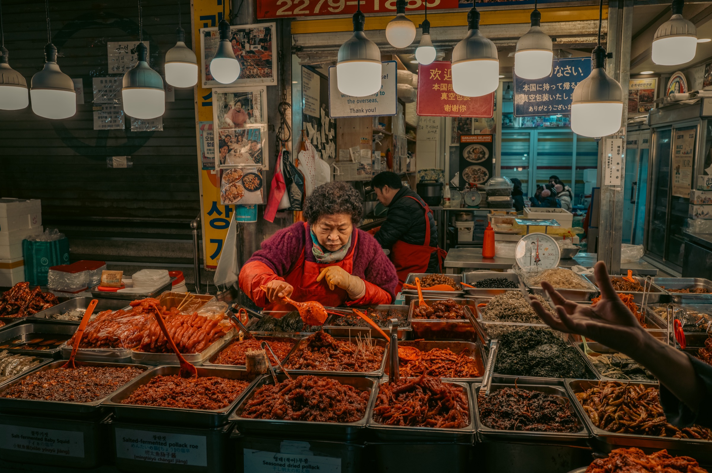 A banchan vendor under a row of bare market bulbs, reaching over trays of pickled sides as a customer gestures