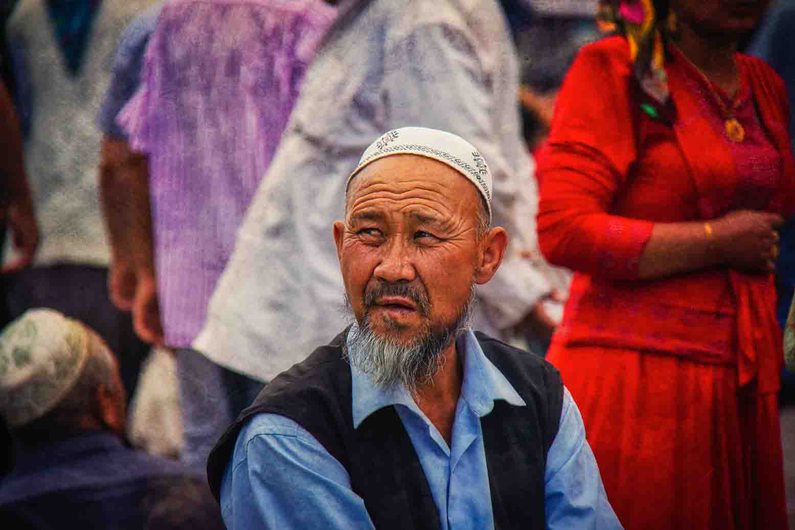 Elder in a white prayer cap and dark vest, a woman in red standing behind