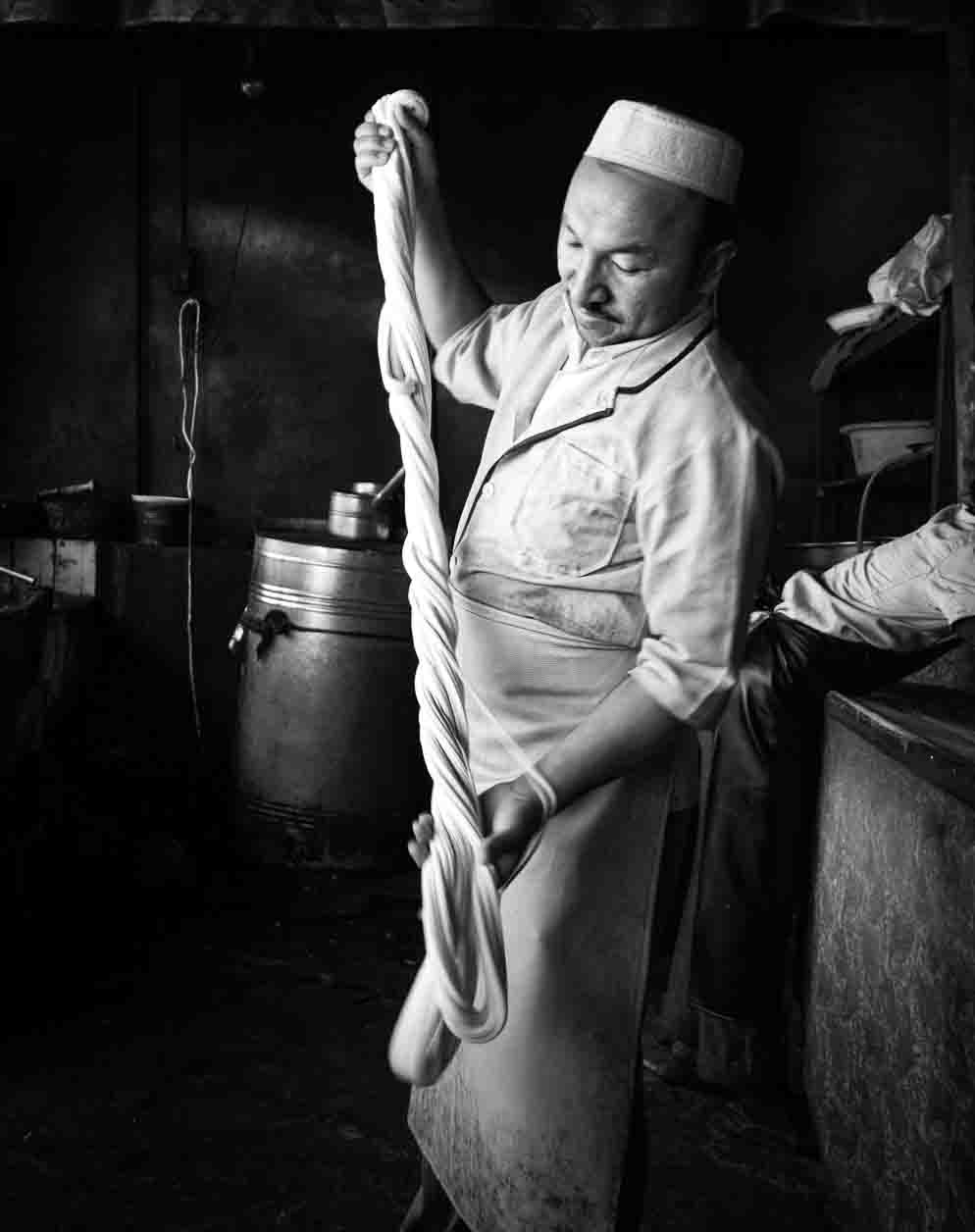 Noodle-puller stretching dough in a dim workshop, in black and white
