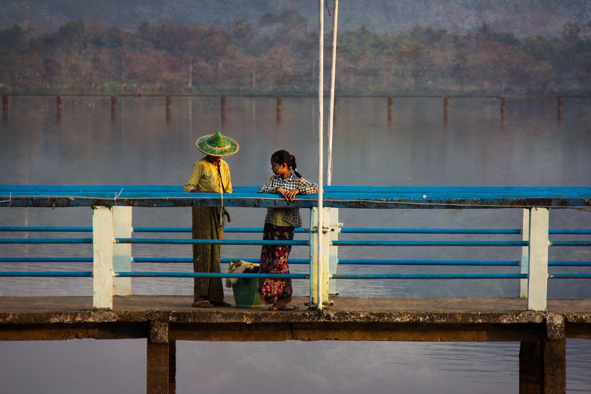 Two women talking at the blue railing of a concrete jetty, one in a conical hat, still lake water and a misted treeline behind them.