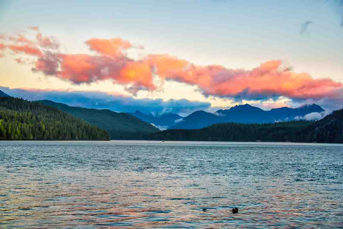 Pink sunset clouds over a forested lake and distant peaks