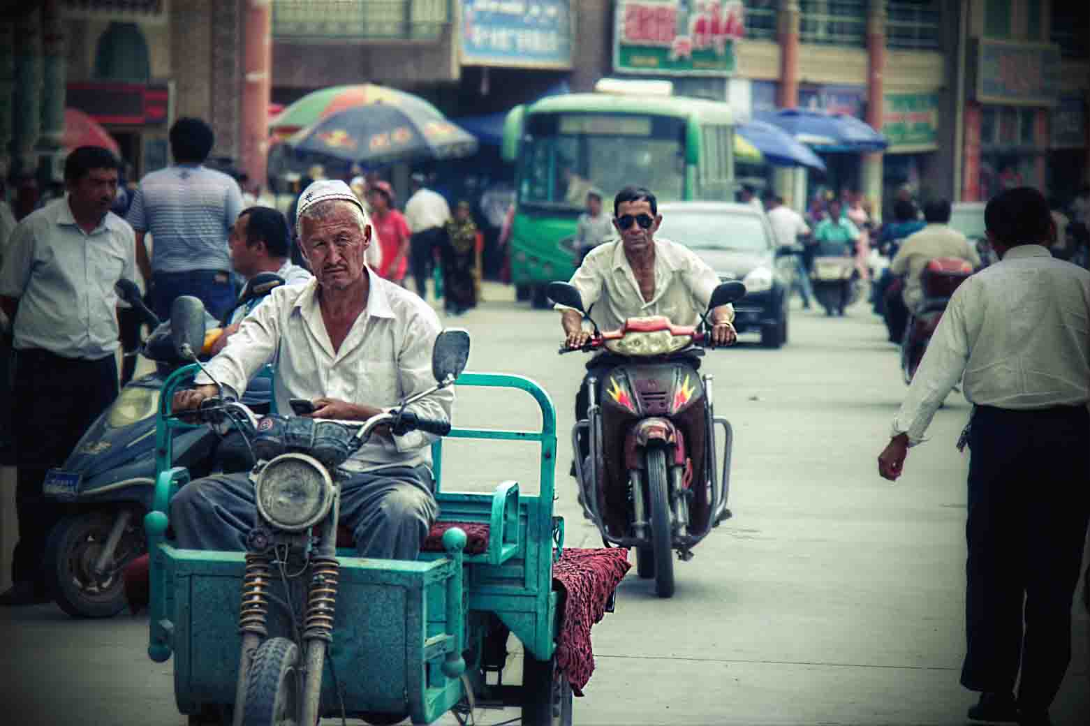 Men riding motorbikes down a Kashgar market street, a green bus behind