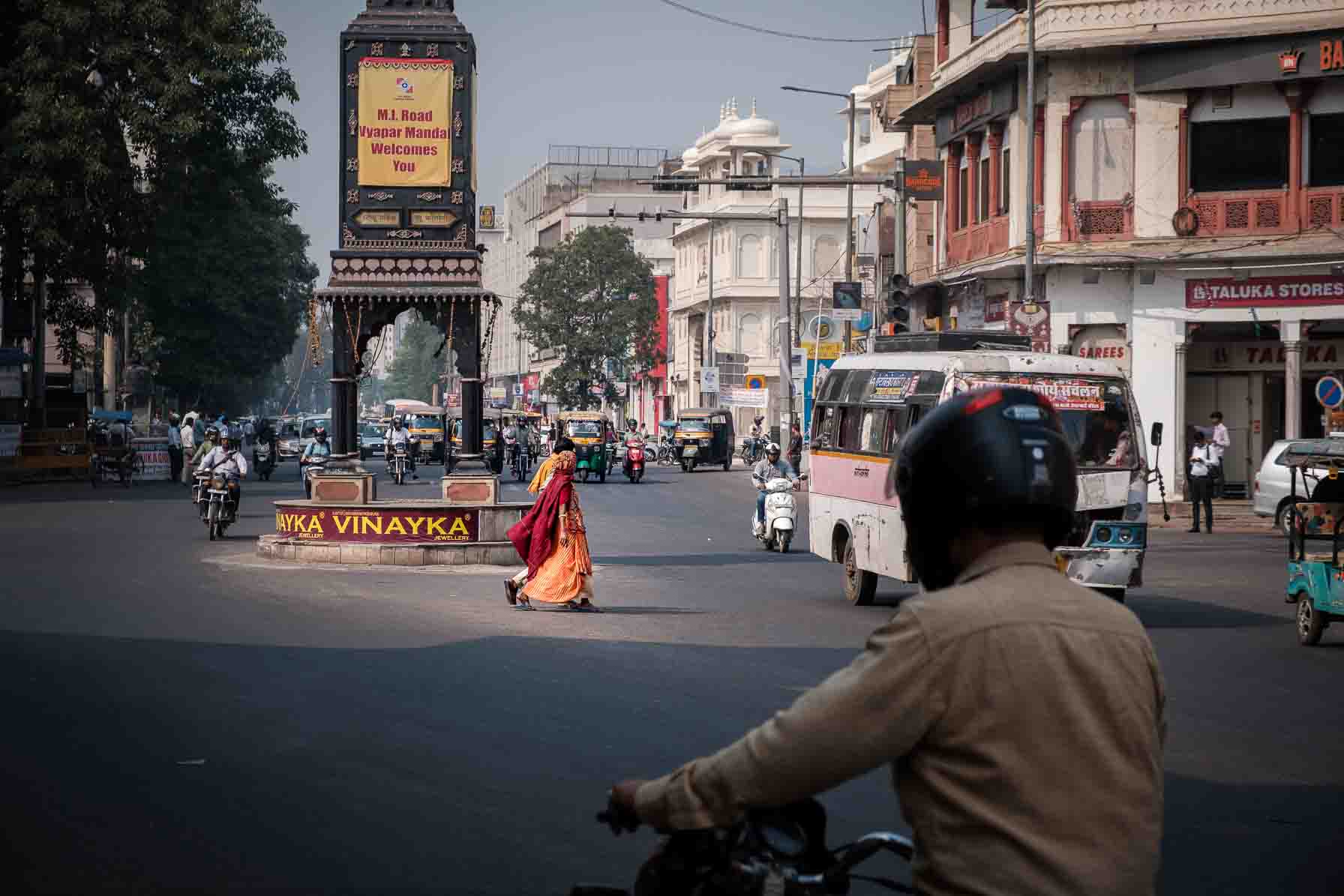 Sari-clad woman crossing a junction with a motorbike rider's back in the foreground